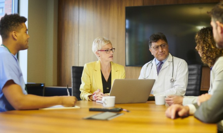 A group of people, including a doctor and nurse, discuss something serious around a boardroom table.
